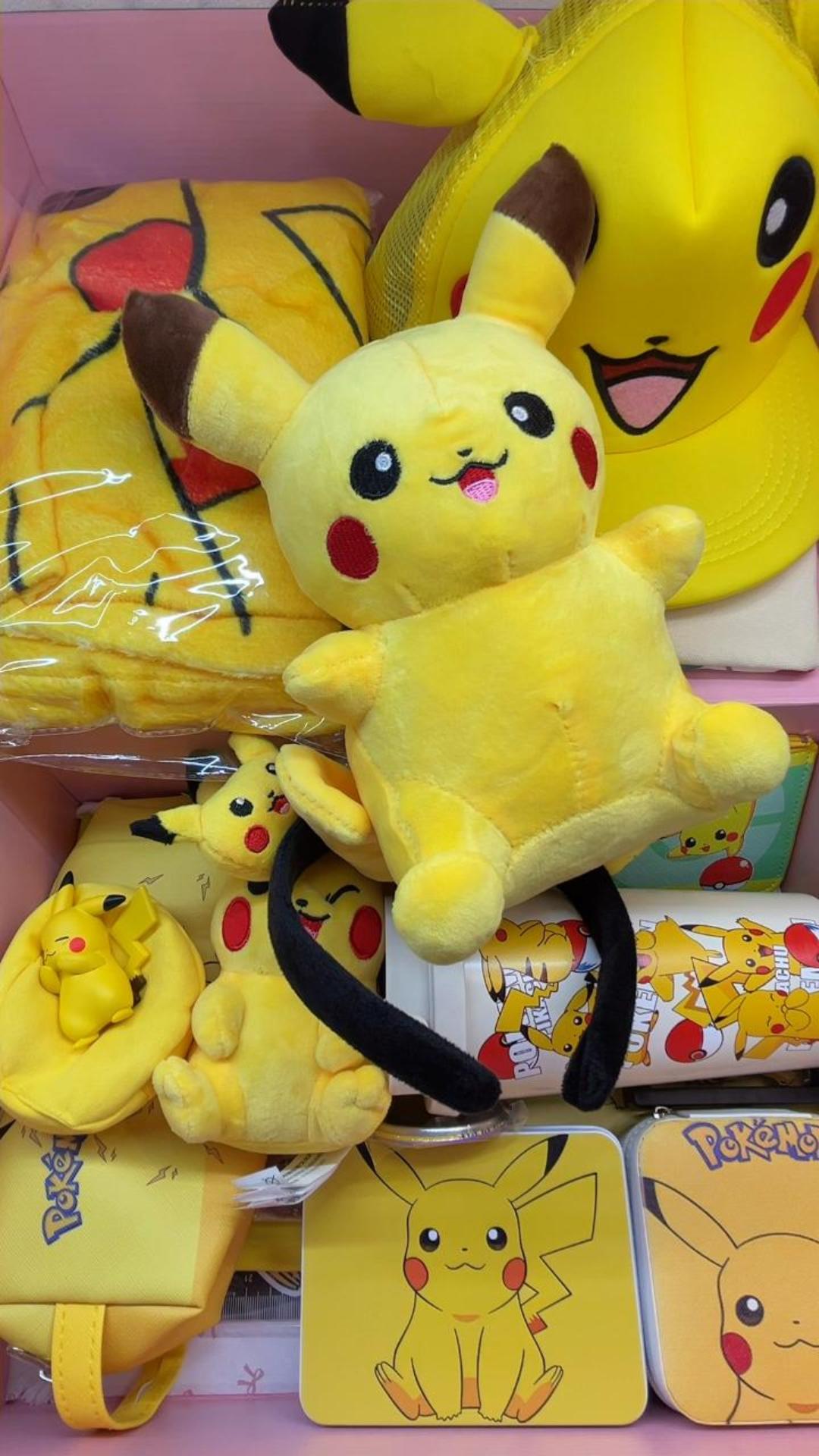 A yellow Pikachu inspired mystery gift box for kids featuring a large character plush toy, vibrant graphic baseball cap, fleece blanket, zippered pencil case, plush ears headband, and character water cup inside a pink surprise birthday package.